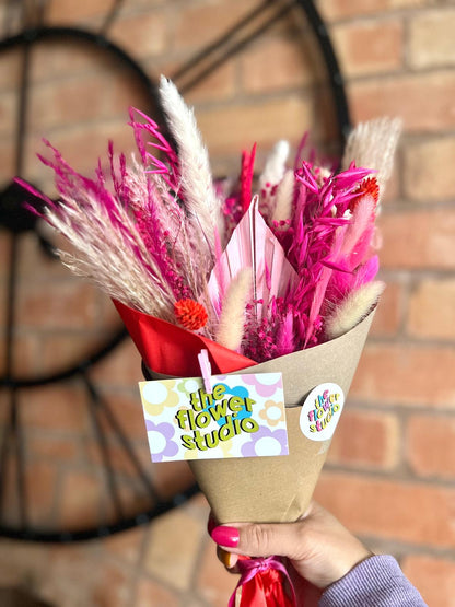 A vibrant bouquet of dried flowers in shades of pink, red, and cream, wrapped in kraft paper with a bold red ribbon. The bouquet is branded with 'The Flower Studio' tags featuring a colourful, retro-inspired design. It is held against a backdrop of a rustic brick wall and a large black clock, adding a modern yet charming touch.