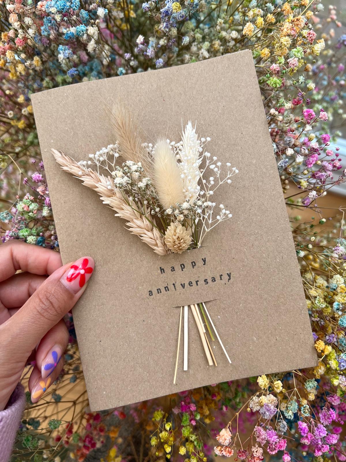 Handmade 'Happy Anniversary' card with dried flowers arranged in a removable slot. The flowers can be taken out and placed in a small vase, creating a lasting keepsake for the special occasion.