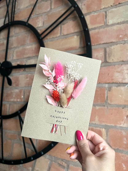 A handmade Valentine’s Day card featuring a delicate arrangement of dried flowers in shades of pink, red, and beige, attached to a kraft card with 'Happy Valentine’s Day' stamped in a minimalist font. The card is held in front of a rustic brick wall and a stylish black clock, creating a warm and elegant aesthetic