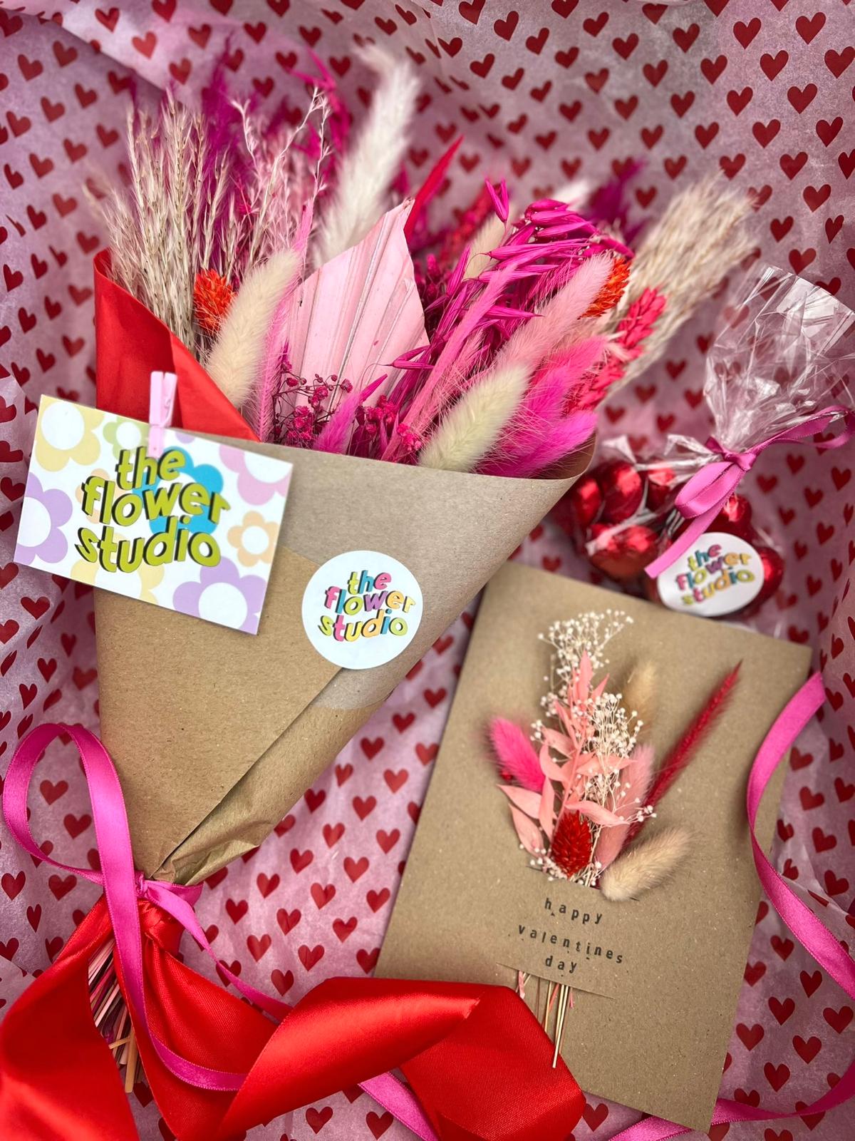 A beautifully arranged Valentine’s Day gift set featuring dried flower bouquets wrapped in kraft paper, tied with vibrant pink and red ribbons, and adorned with 'The Flower Studio' tags. The set also includes a handmade Valentine’s card with dried floral accents and a small bag of heart-shaped chocolates, all displayed on heart-patterned wrapping paper